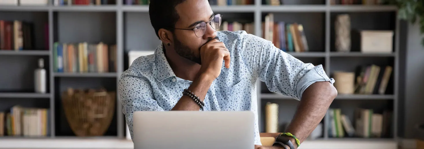 Young black employee with beard wearing glasses sits at his desk, looking off into distance.