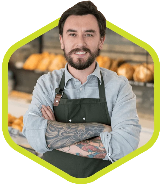 Bearded thirty-something man with tattooed, crossed arms wears an apron and stands in front of baked goods.